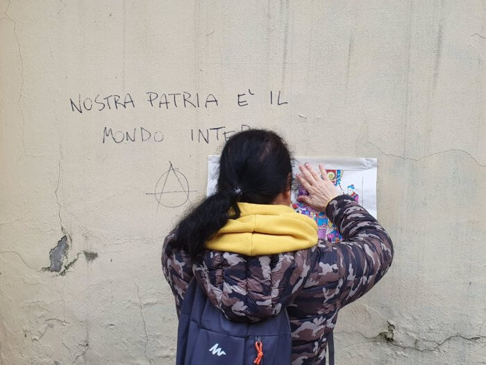 Fig. 4: “Our home is the whole wide world.” A participant in Ai Teng’s intervention posts a nianhua print next to a graffiti with lyrics from the traditional anarchist song “Stornelli d’Esilio” (Pietro Gori, 1895).