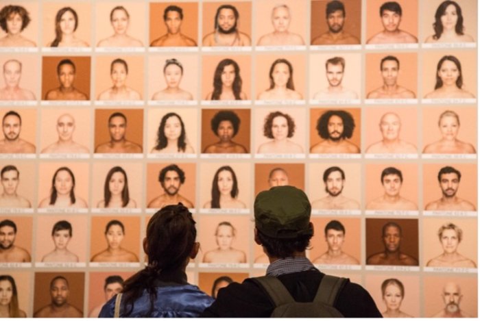 two people standing in front of a screen showing photographs of people of different skin tones, part of the project Humanae